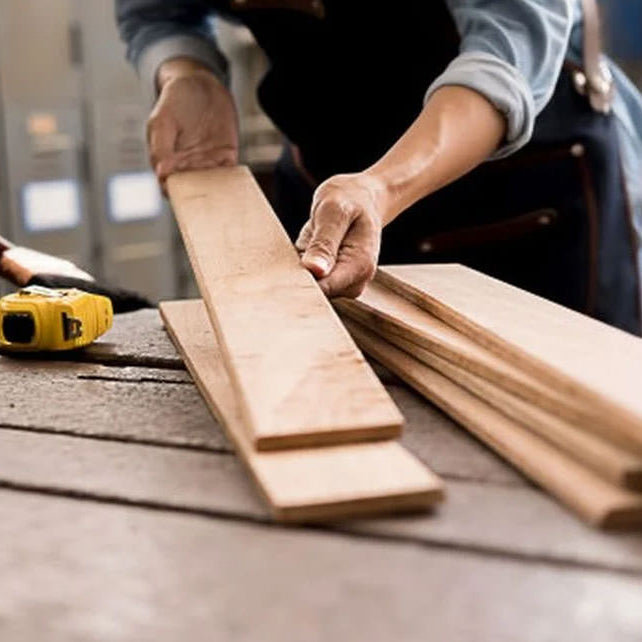 A person in a workshop handling several light-colored wooden boards on a workbench, with tools like a yellow tape measure, glue, and chisels visible in the background.