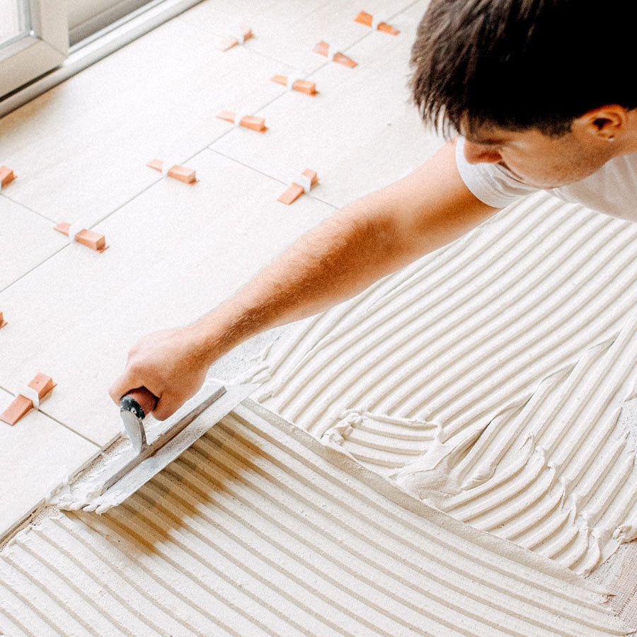 A man applying adhesive or grout with a trowel on a tiled floor, using small orange tile clips to hold the tiles in place near a window.
